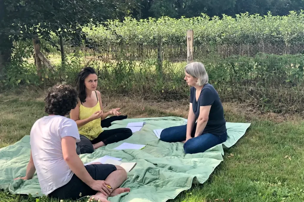 Groupe de trois personnes en discussion lors d'un atelier collaboratif en extérieur, assises sur une couverture dans un cadre naturel.