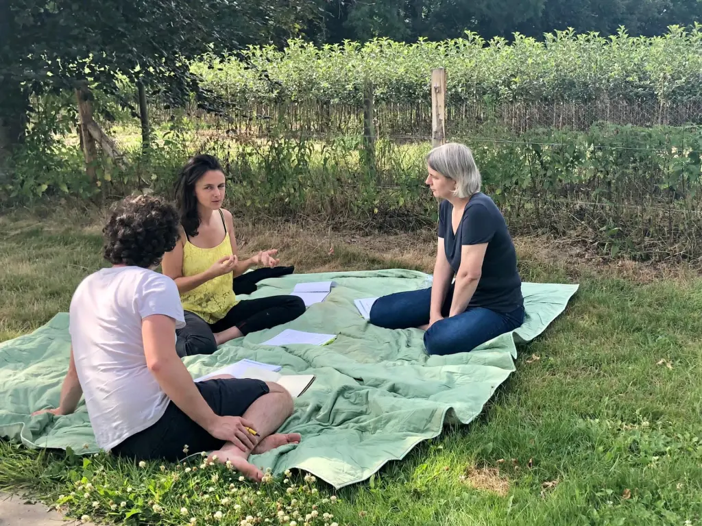 Groupe de trois personnes en discussion lors d'un atelier collaboratif en extérieur, assises sur une couverture dans un cadre naturel.